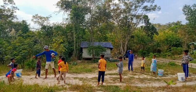 Children and an adult playing a ball game on a dirt clearing, with trees and a small house behind