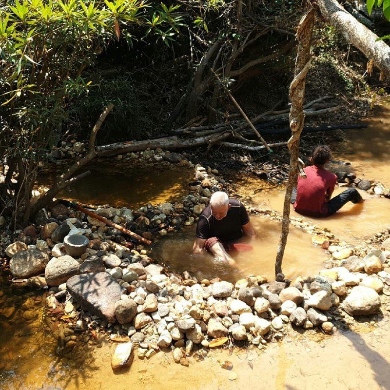 Two people sitting in shallow brown water, moving stones along the edge of the pool